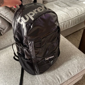 Svart ryggsäck från Supreme - Säljer en svart Supreme ryggsäck med stor logga på framsidan. Väskan har flera dragkedjefack, elastiska snören på framsidan och vadderade axelremmar för extra komfort. Perfekt för att bära med sig allt man behöver till skolan eller stan.