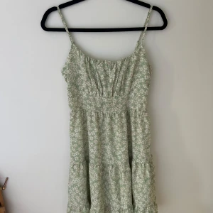 Grön blommig sommarklänning med smock - Söt ljusgrön klänning med vita blommor och smala axelband. Klänningen har smock i ryggen och en lätt rynkad byst. Perfekt för varma dagar och har en luftig passform.