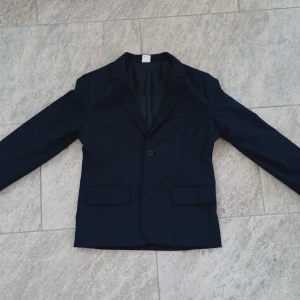 Mörkblå kavaj från Lindex Kids - Snygg mörkblå kavaj från Lindex Kids i klassisk modell med två knappar framtill och fickor med lock. Perfekt för speciella tillfällen som bal eller fest. Passar barn i åldern 9-10 år.