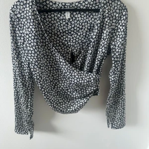 Svart blommig omlottblus från H&M - Säljer en svart omlottblus från H&M med vitt blommönster. Blusen har långa ärmar och en snygg omlottdesign framtill. Perfekt för dig som gillar blommiga mönster och vill ha en feminin look.
