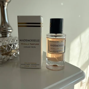 Mademoiselle Eau de Parfum från Morale Parfums Collection - Elegant parfymflaska med ljusrosa doft och svart lock. Förpackningen är vit med guldiga detaljer och svart text. Flaskan är genomskinlig och har en stilren design som passar perfekt på sminkbordet. 