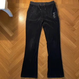 Marinblåa mjukisbyxor från Juicy Couture - Marinblåa mjukisbyxor från Juicy Couture med bootcut-modell och dragsko i midjan. Byxorna har ett glittrigt JC-emblem. Superbekväma och snygga