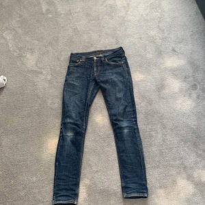 Mörkblå jeansbyxor från Nudie Jeans - Säljer ett par mörkblå jeans från Nudie Jeans med klassisk femficksdesign och orangea sömmar. Byxorna har smal passform och raka ben, perfekt för en stilren vardagslook.