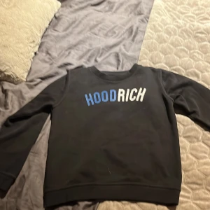 Hoodrich tröja  - Säljer en svart sweatshirt från Hoodrich i storlek M (10-12 år). Tröjan har tryckt text 'HOODRICH' i blått och vitt framtill och är tillverkad i mjukt material. Klassisk rund halsringning och långa ärmar.