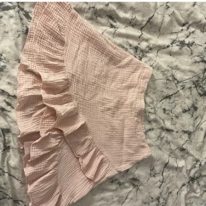 Ljusrosa volangkjol - Supersöt ljusrosa kjol med volangdetalj längs ena sidan. Kjolen är kort och har en luftig, vågig struktur i ett mjukt tyg. Perfekt för varma dagar och enkel att matcha med olika toppar. Dock liten i storlek, köpt för 450kr