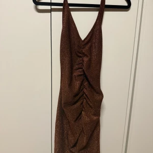 Brun glittrig omlottklänning - Säljer en brun glittrig klänning med omlottdetalj och rynk framtill. Klänningen är ärmlös med djup v-ringning och smala axelband. Perfekt för dig som vill sticka ut med en snygg och trendig look.