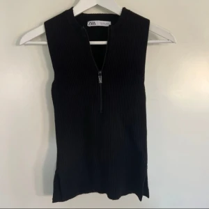 Svart ribbad topp  från Zara - Säljer en svart, ärmlös ribbad topp från Zara med dragkedja framtill. Toppen har en tight passform och är tillverkad i ett stretchigt material som sitter snyggt på kroppen. Perfekt för dig som gillar minimalistisk stil. 