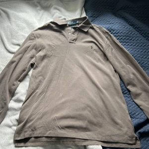 Brun långärmad pikétröja från Polo Ralph Lauren - Säljer en klassisk brun långärmad pikétröja från Polo Ralph Lauren med krage och knappar framtill. Tröjan har broderad logga på bröstet och är tillverkad i bomull. Perfekt för en stilren och avslappnad look.
