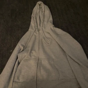 Grå hoodie med dragkedja från H&M Divided - Grå hoodie från H&M Divided med dragkedja framtill och huva med dragsko. Tröjan har två fickor på magen och är långärmad. Perfekt för en avslappnad stil.