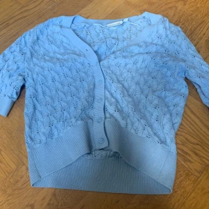 Ljusblå stickad kofta från Kappahl - Säljer en ljusblå stickad kofta från Kappahl med vackert hålmönster och knappar framtill. Koftan har trekvartsärm och bred ribbad kant nedtill och vid ärmslut. Perfekt för lager-på-lager och en söt detalj till din outfit.