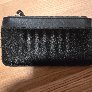 Svart plånbok med pälsdetalj - Säljer en svart plånbok med dragkedja och handledsrem. Framsidan har en mjuk pälsliknande detalj som ger en lyxig känsla. Plånboken har flera fack inuti för kort och sedlar. Perfekt accessoar för att hålla ordning på dina värdesaker.