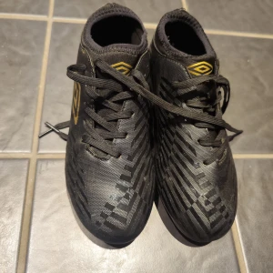 Svarta fotbollsskor från Umbro - Säljer ett par svarta fotbollsskor från Umbro med gulddetaljer och snörning. Skorna har ett diskret mönster och dobbar för spel på gräs. Perfekta för dig som vill ha stilrena och funktionella skor på planen.