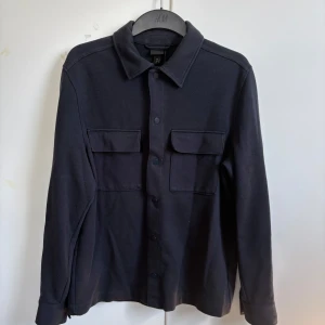 Marinblå overshirt - Stilren marinblå overshirt från H&M med klassisk krage och två stora bröstfickor med lock. Skjortan har knäppning framtill och långa ärmar, perfekt att bära som lager över en t-shirt.