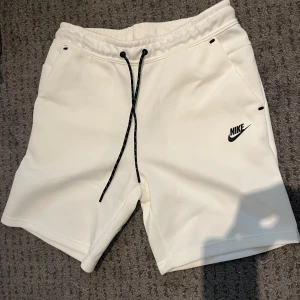 Vita nike tech shorts - Säljer ett par vita Nike shorts med svart logga på vänster ben. Shortsen har snörning i midjan och dragkedjeficka bak. Perfekta för träning eller chill. Mjukt och stretchigt material.