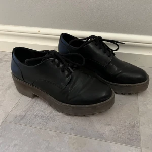 Svarta platåskor med snörning - Säljer ett par svarta platåskor i skinnimitation med snörning. Skorna har rund tå och en rejäl platt sula som ger extra höjd. Perfekta för dig som vill ha en stilren men edgy look.