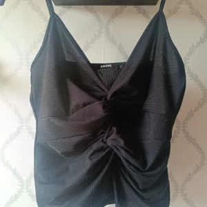 Svart topp med knutdetaljer från BikBok - Säljer en svart topp från BikBok i glansigt, plissera material. Toppen har smala axelband som går att ändra och två snygga knutdetaljer framtill som ger en unik look. Perfekt att styla med jeans eller kjol. Väldigt snygg men kommer inte tills användning, 40 kr 