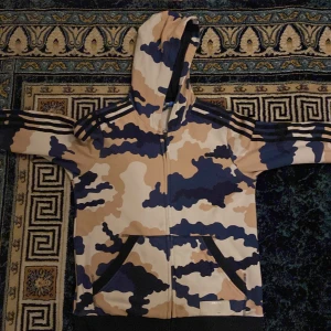 Beige och blå camo hoodie från Adidas - Säljer en hoodie från Adidas med beige, blå och mörkblå camouflage-mönster. Jackan har luva, dragkedja framtill, två fickor och de klassiska Adidas-ränderna på ärmarna samt stor logga på ryggen köpt från Frankrike köpt för 50 euro . Och det skulle uppskattas om personen som är intresserad och vill köpa denna tröja komma hit till Rinkeby området. Om du vill kontakta mig eller swisha här är numret:0769313560