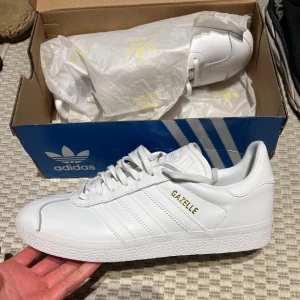 Vita Adidas Gazelle sneakers - Säljer ett par helt vita Adidas Gazelle sneakers i skinn med klassisk design och guldfärgad Gazelle-text på sidan. Skorna har snörning och platt sula, perfekt för dig som gillar stilrena och tidlösa sneakers.