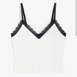 Vit linnetopp med svarta spetsdetaljer - Snygg vit linnetopp med tunna svarta axelband och dekorativ svart spets längs urringningen. Perfekt att styla till jeans eller kjol för en trendig look.