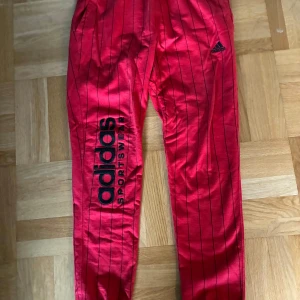 Röda adidas byxor med svarta ränder - Säljer ett par röda adidas byxor med svarta tunna vertikala ränder och stor adidas-logga på ena benet. Byxorna har resår i midjan och en bakficka. Perfekta för en sportig look.
