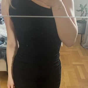 Svart one shoulder klänning - Säljer en svart, tight one shoulder klänning med rynkade detaljer längs sidorna. Klänningen har en asymmetrisk axel och är figurnära, vilket ger en snygg siluett. Perfekt för dig som gillar stilrena och trendiga plagg.