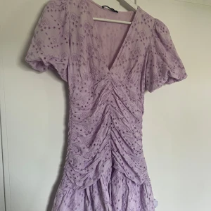 Ljuslila broderad klänning med volang - Superfin ljuslila klänning med v-ringning och korta puffärmar. Klänningen har broderade detaljer och volanger samt rynk framtill för en extra snygg siluett. Perfekt för sommarens alla tillfällen!