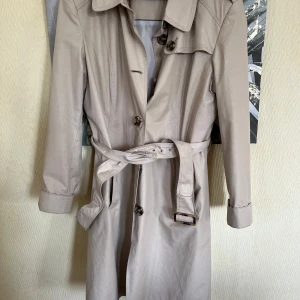 Beige trenchcoat med bälte - Stilren beige trenchcoat med klassisk krage, knappar framtill och tillhörande bälte i midjan. Jackan har långa ärmar med spänne vid ärmslut och är tillverkad i ett bomullsliknande material. Perfekt för övergångssäsonger.