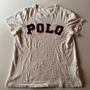 Vit t-shirt från Ralph Lauren - Säljer en vit t-shirt från Ralph Lauren med texten 'POLO' i blått och rött på framsidan. T-shirten är kortärmad och tillverkad i bomull.