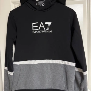 EA7 Emporio Armani hoodie svart/grå - Snygg hoodie från EA7 Emporio Armani i svart, grått och vitt. Tröjan har huva, lång ärm och stor logga framtill. Färgblockad design med breda ränder och känguruficka. Perfekt för dig som gillar streetwear och vill sticka ut med stil.