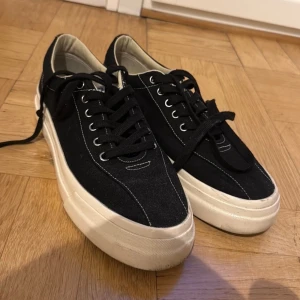 Svarta S.W.C sneakers med vit sula - Svarta sneakers från S.W.C med vit, tjock sula och kontrasterande vita sömmar. Klassisk låg modell med snörning och rund tå. Ovandel i canvas och detaljer i vitt. Perfekta för en clean och stilren look.