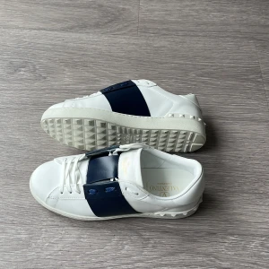 Valentino Garavani Open Navy  - Snygga Valentino Garavani Open sneakers i vitt skinn med bred marinblå läderdetalj över mitten. Klassisk rund tå, vita snören och coola nitar på hälen. Box och påse finns med och skorna är väldigt fräscha, perfekt för dig som vill ha en clean stil