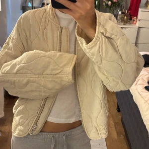 Beige quiltad bomberjacka - Säljer en beige quiltad bomberjacka med oversized passform. Jackan har vågiga sömmar, dragkedja framtill och elastisk nederkant. Perfekt för dig som gillar en chill och trendig look. Passar till allt och är superenkel att styla.