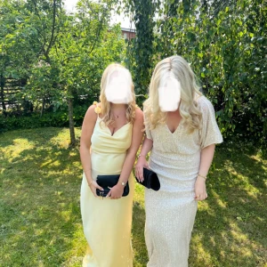 Ljusgul långklänning med blommor - Superfin ljusgul långklänning i satinliknande material med smala axelband och dekorativa blommor längs banden. Klänningen har en snygg draperad urringning och figurnära passform. Perfekt för sommarens speciella tillfällen!