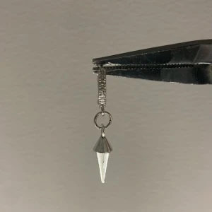 Silvrigt örhänge med konformad detalj - Säljer ett stilrent örhänge i silverfärg med en konformad hängande detalj. Örhänget har minimalistisk design och passar dig som gillar edgy och modern stil. Perfekt att mixa med andra smycken för en personlig look.