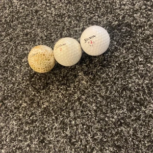 3 golfbollar 5 kronor styck 15 kronor för alla - Bilderna visar tre golfbollar av märkena Titleist och Srixon, placerade på en mörk, mönstrad matta. En av bollarna är tydligt använd och missfärgad, medan de andra två är i bättre skick. Perfekt för dig som gillar golf och vill ha extra bollar till träning eller spel.