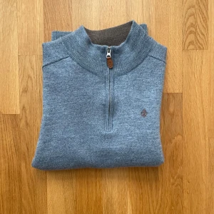 Blå merinoulltröja från Morris half zip - Snygg blå tröja från Morris i 100% merinoull med half zip och brunt innerkrage. Tröjan har lång ärm, ribbade muddar och en liten broderad logga på bröstet. Perfekt för dig som gillar stilren och bekväm look. Tröjan är i storlek Medium men har krympt i tvätten till storlek XS-S och blivit väldigt slim fit, matrialet har dessutom blivit urtvättat.