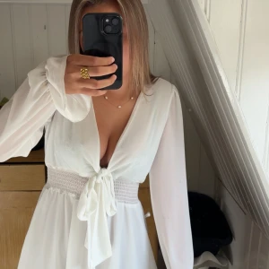 Vit kort omlottklänning med volang - Supersöt vit kortklänning med omlottknytning framtill, långa transparenta ärmar och smock i midjan. Klänningen har v-ringning och volangdetaljer längst ner. Perfekt för sommarens alla tillfällen! Endast använd en gång! 
