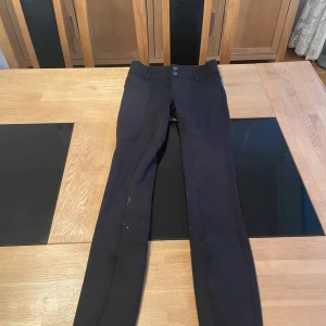 Svarta ridbyxor med silikonprint - Svarta ridbyxor från kingston i stretchigt material med silikonprint på insidan av benen för extra grepp. Byxorna har hög midja, två knappar i midjan och är tillverkade i 90% polyester och 10% elastan. Snygg och tight passform som sitter på plats när du rider. De kan dock vara lite tighta i låren o smalbenen så tänk på det innan du säger att du är intresserad! Pris kan diskuteras vid snabb affär.