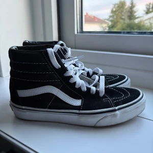 Svarta Vans Sk8-Hi sneakers - Klassiska Vans Sk8-Hi sneakers i svart med vit sula och vit sidosnirk. Högt skaft, vita snören och tydliga kontrastsömmar. Ovandel i mocka och canvas för en skön och cool streetstil.