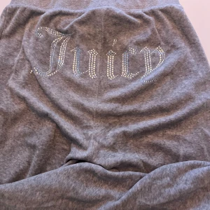 Grå Juicy Couture mjukisbyxor med strass - Grå mjukisbyxor från Juicy Couture med glittrande strassdetaljer på bakfickan där det står 'Juicy' och ett litet J på framsidan. Byxorna har dragsko i midjan och är tillverkade i mjukt material för maximal komfort.