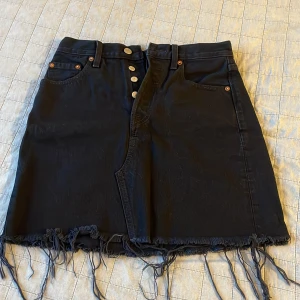 Svart jeanskjol från Levi’s - Svart jeanskjol från Levi's med slitningar nere. Klassiska femficksdetaljer och bälteshällor. Storleken på kjolen är 26 (skulle säga att det är xs/s). Färgen är typ urtvättad svart.