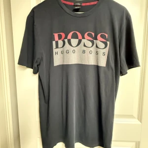 Svart Hugo Boss t-shirt med tryck - Svart t-shirt från Hugo Boss i bomull med stort BOSS-tryck i rött och grått framtill. Klassisk rund halsringning och korta ärmar. Snygg och enkel design som passar till det mesta.