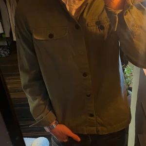 Grön overshirt med knappar - Snygg militärgrön overshirt med klassisk krage och svarta knappar framtill. Två bröstfickor med lock och knappdetaljer ger en cool utility-vibe. Perfekt att slänga över en t-shirt för en avslappnad look.