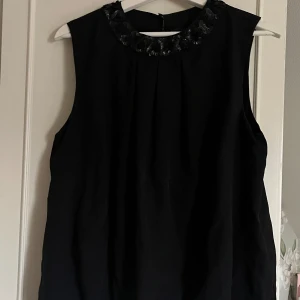 Svart ärmlös topp med paljetter - Snygg svart ärmlös topp med rund halsringning dekorerad med svarta paljetter. Perfekt för dig som vill ha en stilren men ändå festlig look. Lätt och luftig modell som passar till många olika outfits. 