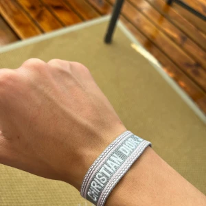 Grått Christian Dior J'Adior armband - Snyggt armband från Christian Dior med texten 'J'Adior' broderad i vitt på grå bakgrund. Armbandet är gjort i tyg och har en stilren, minimalistisk look som passar perfekt till din outfit. Justerbar knytning för bästa passform.