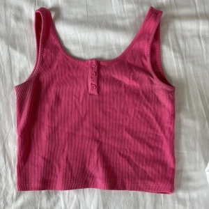 Rosa ribbad linnetopp med knappar från Gina - Köpt på Gina Tricot (barnavdelningen). Inga defekter. Storlek 152, men passar XXS/XS. Superfin rosa linnetopp i ribbad bomull med tre knappar framtill. Croppad modell med breda axelband och rundad halsringning. Perfekt för varma dagar och enkel att matcha med jeans eller kjol.