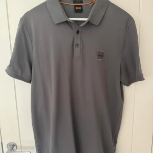 Grå pikétröja från BOSS slim fit - Snygg grå pikétröja från BOSS i slim fit-modell. Klassisk krage, korta ärmar och knäppning framtill med tre knappar. Diskret BOSS-logga på bröstet. Perfekt för en clean och stilren look.