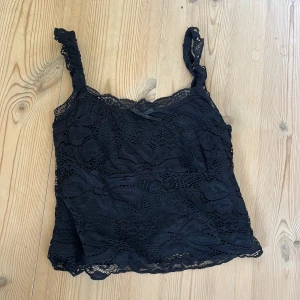 Svart spetslinne med rosett - Säljer ett svart linne i spets med smala axelband och en liten söt rosett framtill. Toppen har ett genombrutet spetsmönster som ger en romantisk vibe. Jättesöt men aldrig använd då den var lite kort på mig. Tvättad 1 gång. ❤️