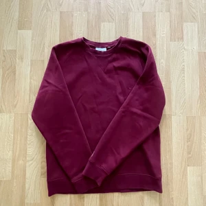 Vinröd sweatshirt från Carhartt - Vinröd sweatshirt från Carhartt med klassisk rund halsringning och ribbade muddar. Tröjan har en diskret Carhartt-logga i guld på ärmen och är tillverkad i mjuk bomull. Perfekt för dig som gillar enkel och stilren streetwear.