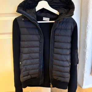 Svart Moncler pufferjacka med huva - Svart pufferjacka från Moncler med stickade ärmar och vadderad front. Jackan har huva, dragkedja och Moncler-logga på ärmen. Perfekt för dig som vill ha en snygg och sportig look med premiumkänsla. Kan sänka vid snabb affär 